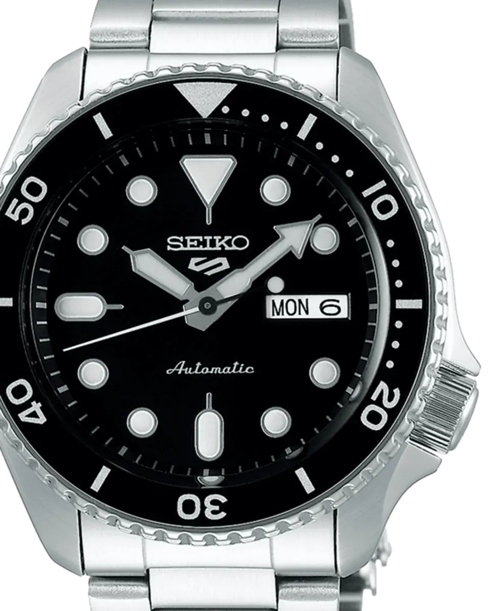 Primer plano frontal de la esfera negra, el bisel rotatorio y la corona del reloj sobre fondo blanco.