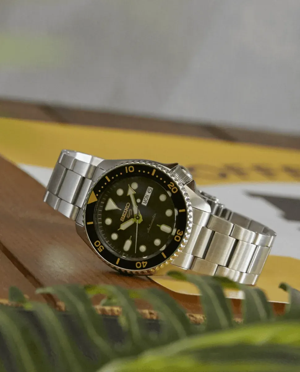 Reloj visto en perspectiva diagonal sobre superficie de madera, con hojas verdes desenfocadas en primer plano.