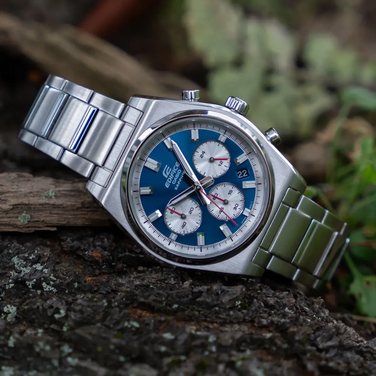 Reloj apoyado diagonalmente sobre corteza de árbol oscura, fondo natural desenfocado.