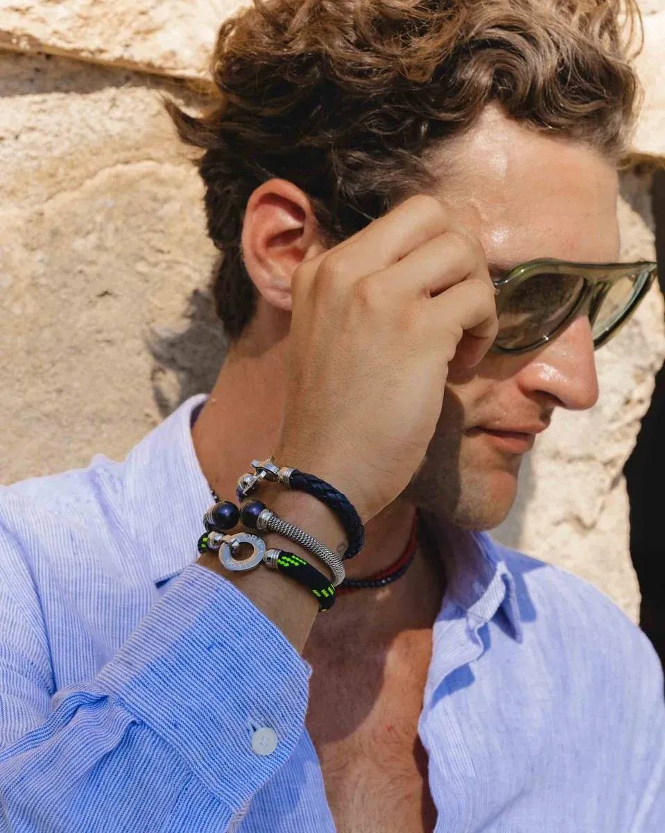 Close-up of wrist wearing blue braided and steel bracelet, model's hand near sunglasses.