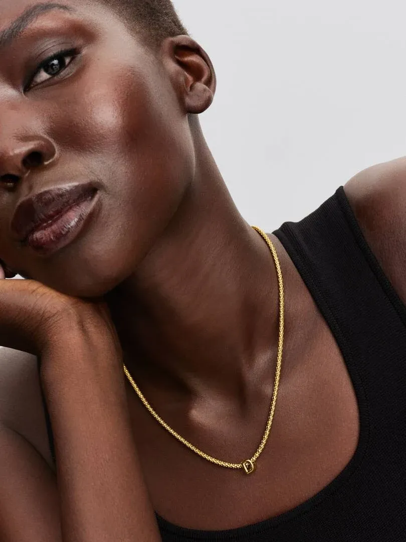 Primer plano de collar con letra D sobre cuello y pecho, modelo con top negro, fondo neutro.