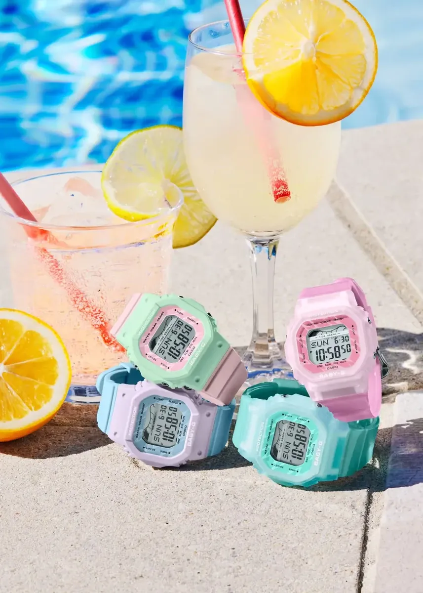 Bodegón de cuatro relojes digitales en tonos pastel sobre piedra, con copas de limonada y piscina desenfocada al fondo.