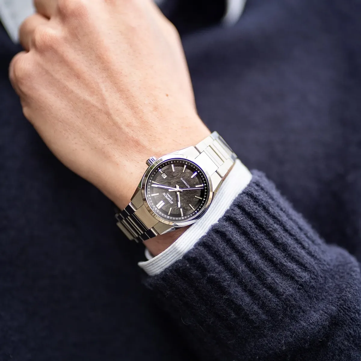 Reloj puesto en la muñeca, primer plano con jersey azul marino y puño de camisa blanca.