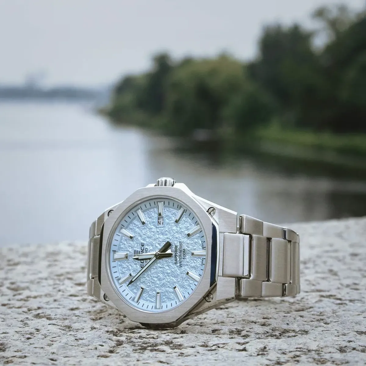 Reloj apoyado sobre superficie de piedra, toma en ángulo tres cuartos con fondo natural desenfocado de río y vegetación.