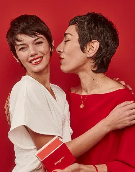 Twee vrouwen die elkaar omhelzen tegen een rode achtergrond, een draagt de gouden medaillonketting.