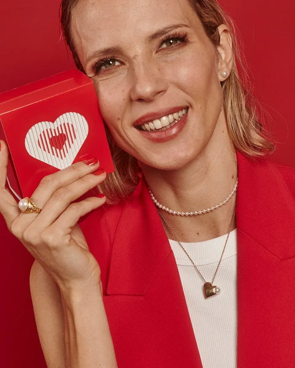 Mujer sonriente con collar de corazón y perla, fondo rojo vibrante, plano medio corto.