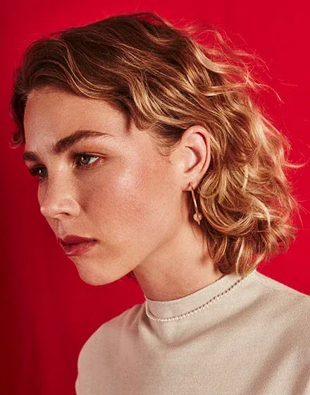 Primer plano de perfil con collar de perlas y pendiente dorado sobre modelo, fondo rojo intenso.
