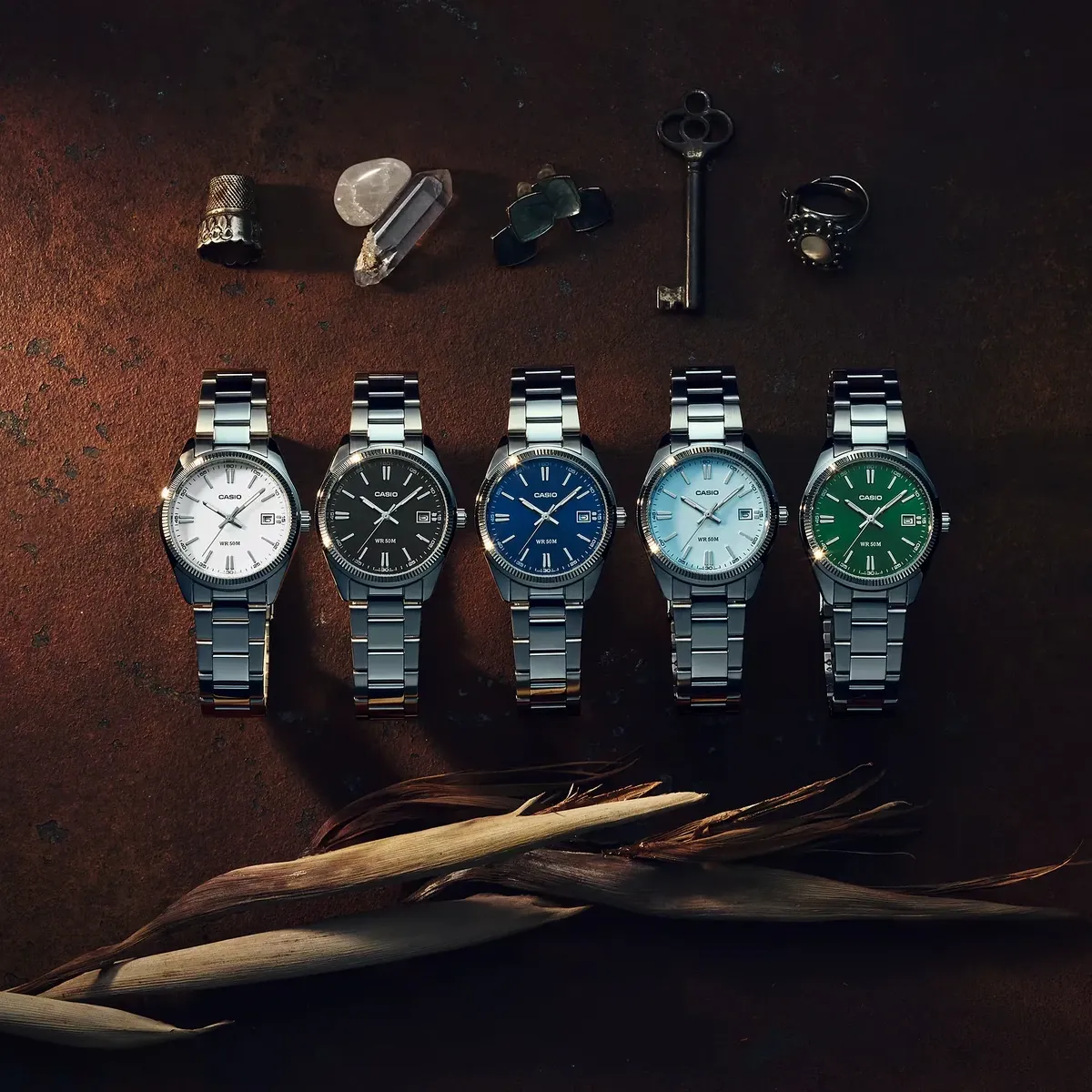 Vista cenital de cinco relojes de acero con esferas de distintos colores y elementos decorativos sobre fondo marrón.