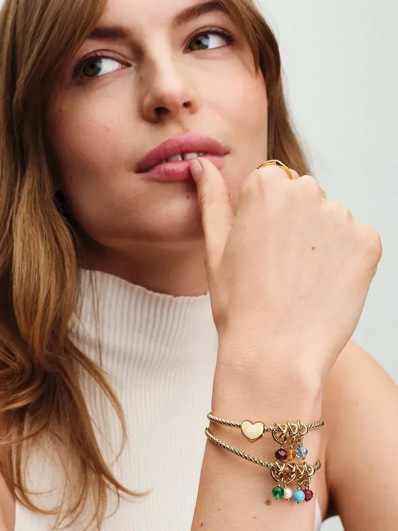 Close-up van pols met gouden armband en charms, model in witte coltrui, wazige achtergrond.