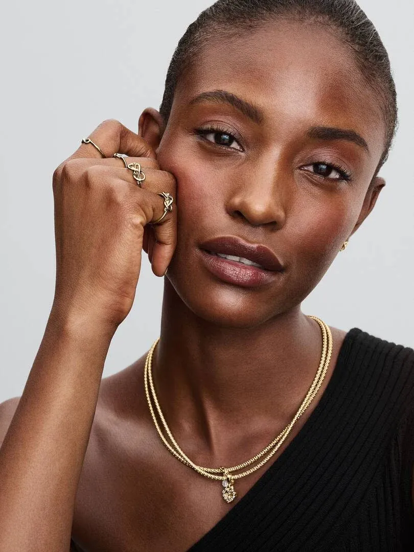 Close-up of model wearing double heart necklace and rings, light neutral background.