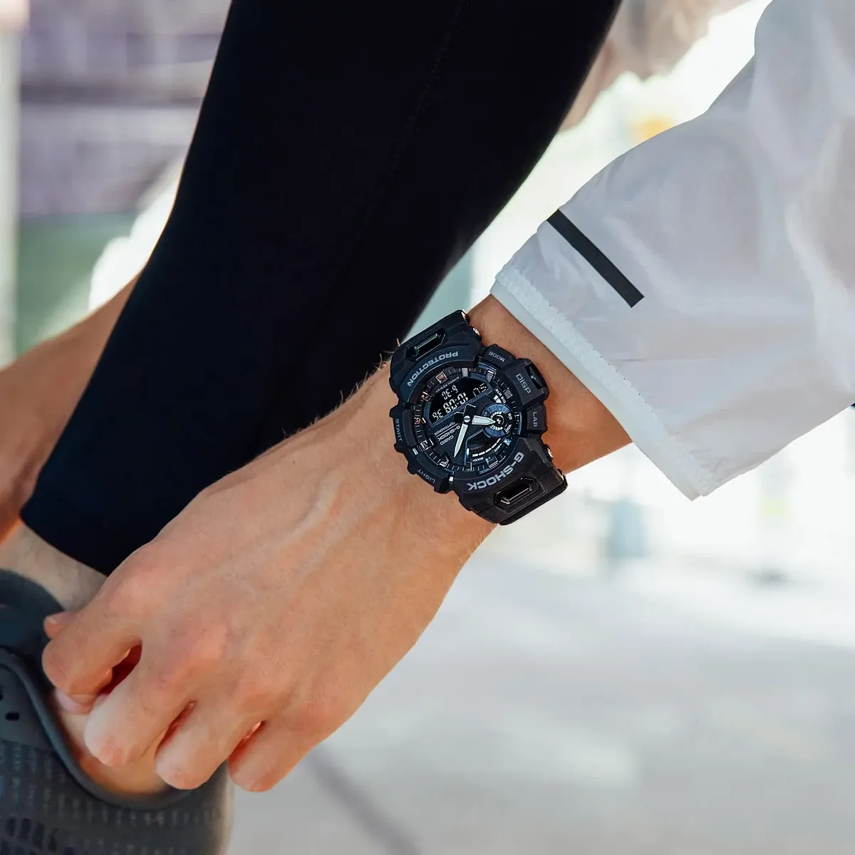 Reloj puesto en la muñeca de un modelo con ropa deportiva, primer plano, fondo urbano desenfocado.