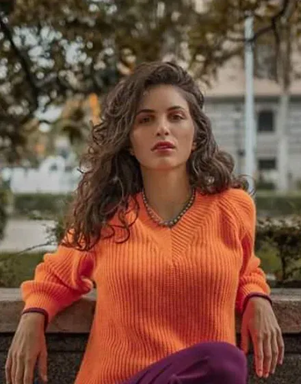 Modelo con jersey naranja luciendo el collar de perlas, toma frontal de medio cuerpo en exterior desenfocado.