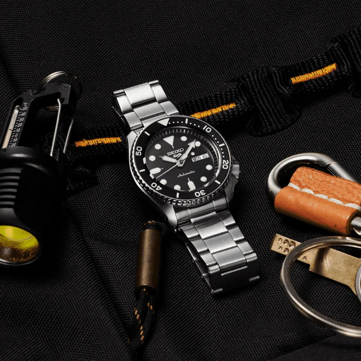 Vista cenital del reloj sobre fondo de tela negra, rodeado de accesorios como una linterna y un llavero de cuero.