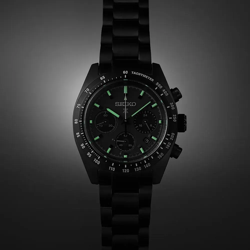 Vista frontal del reloj con índices y agujas luminiscentes, centrado sobre fondo gris oscuro neutro.