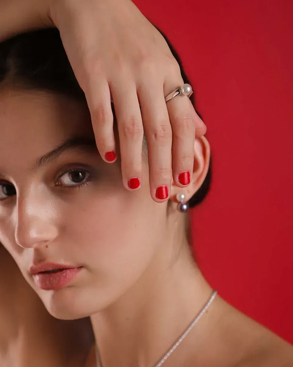 Primer plano de mano con anillo de perla sobre el rostro, fondo rojo sólido y uñas lacadas en rojo.