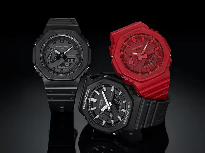 Composición de tres relojes, uno rojo y dos negros, sobre superficie negra reflectante y fondo negro.