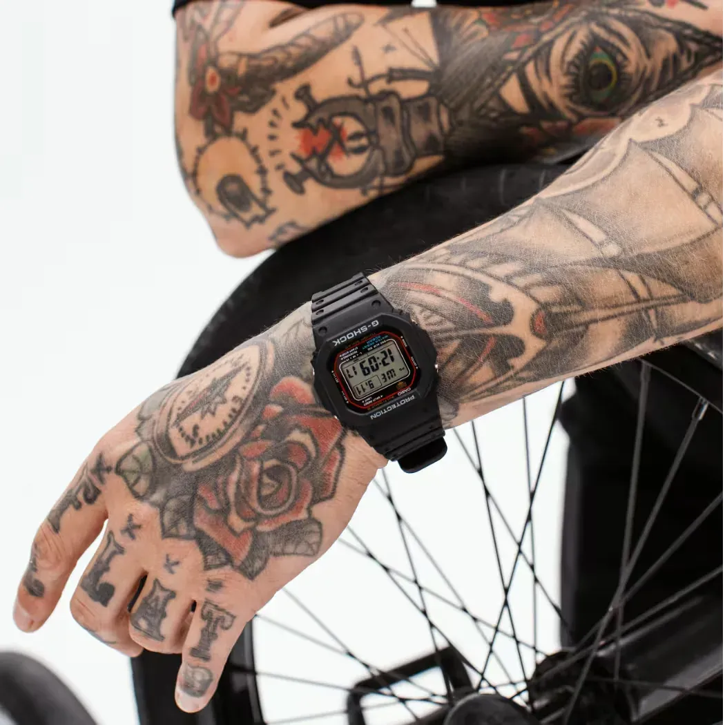 Reloj puesto en muñeca tatuada, apoyada sobre la rueda de una bicicleta, fondo blanco.