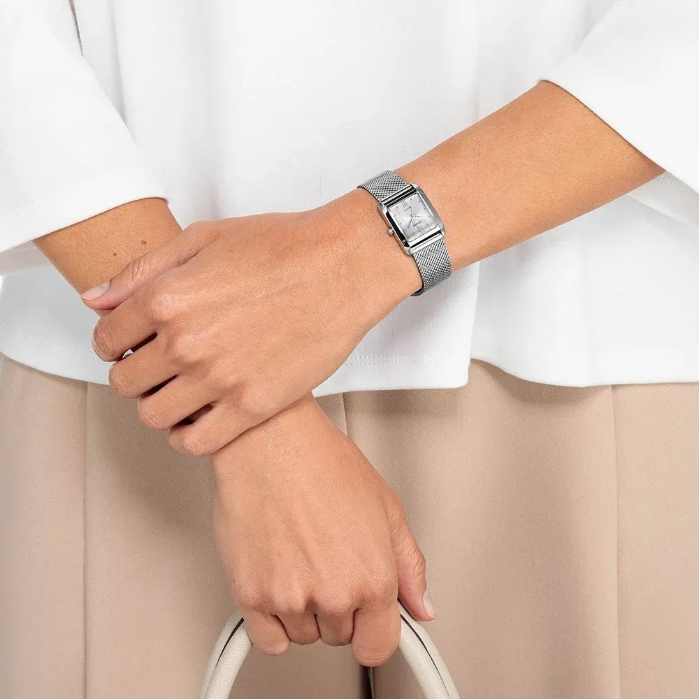 Watch worn on a model's wrist, framing of hands over white clothing and a beige skirt.