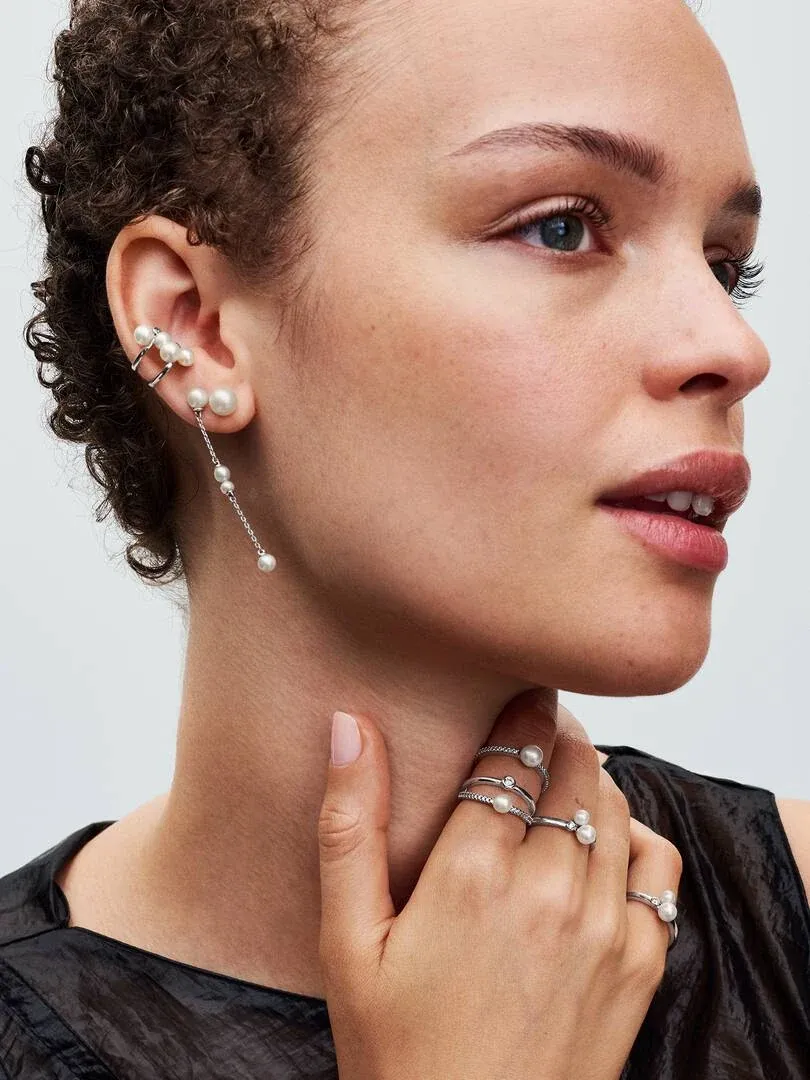 Primer plano de perfil con modelo luciendo pendientes y anillos de perlas, fondo neutro claro.