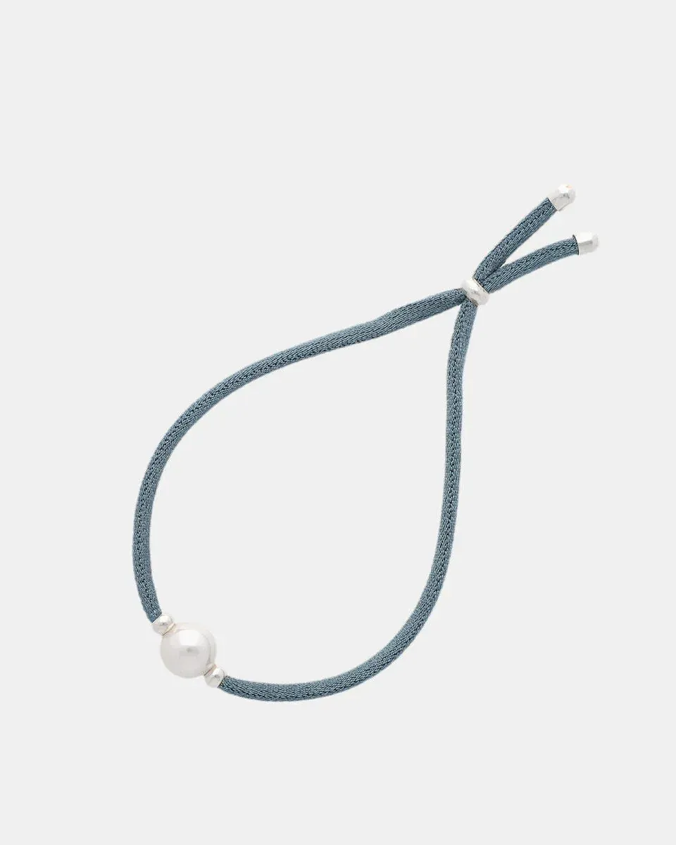Pulsera de cordón azul con una perla blanca central, vista cenital sobre fondo gris claro.