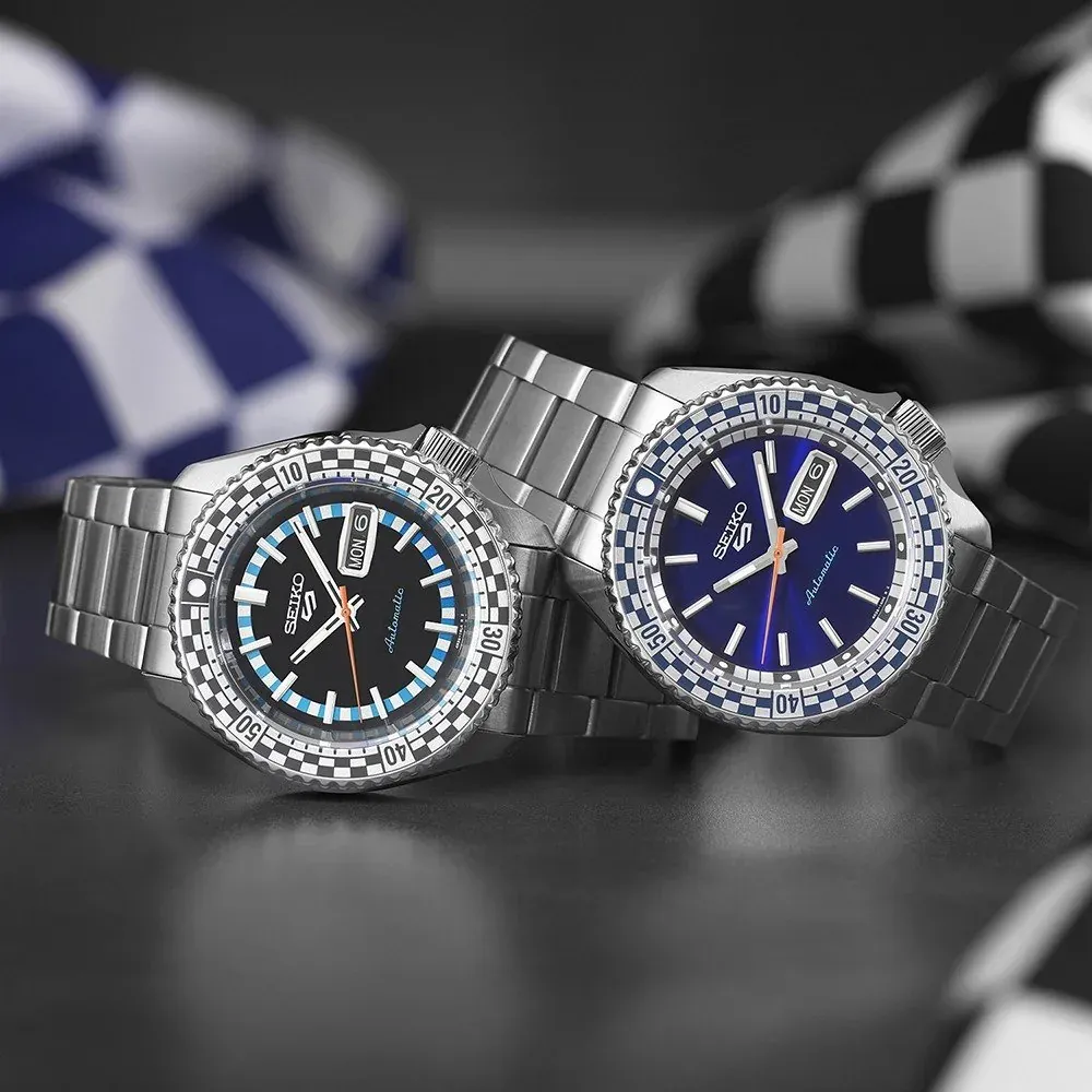 Two watches arranged on a dark surface with blurred checkered flags in the background.