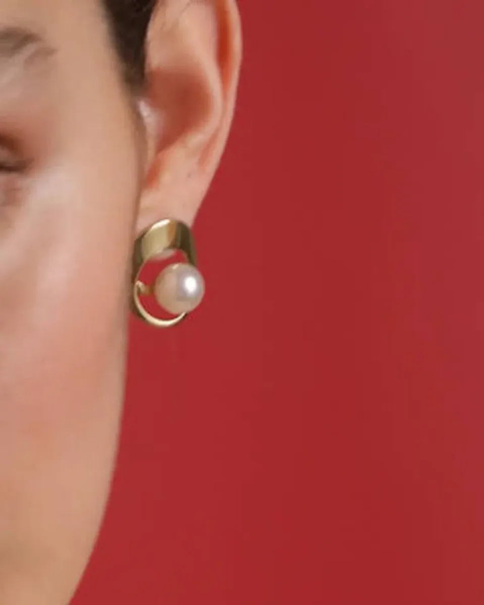 Gros plan latéral d'une boucle d'oreille en perle blanche et structure dorée portée à l'oreille, fond rouge.
