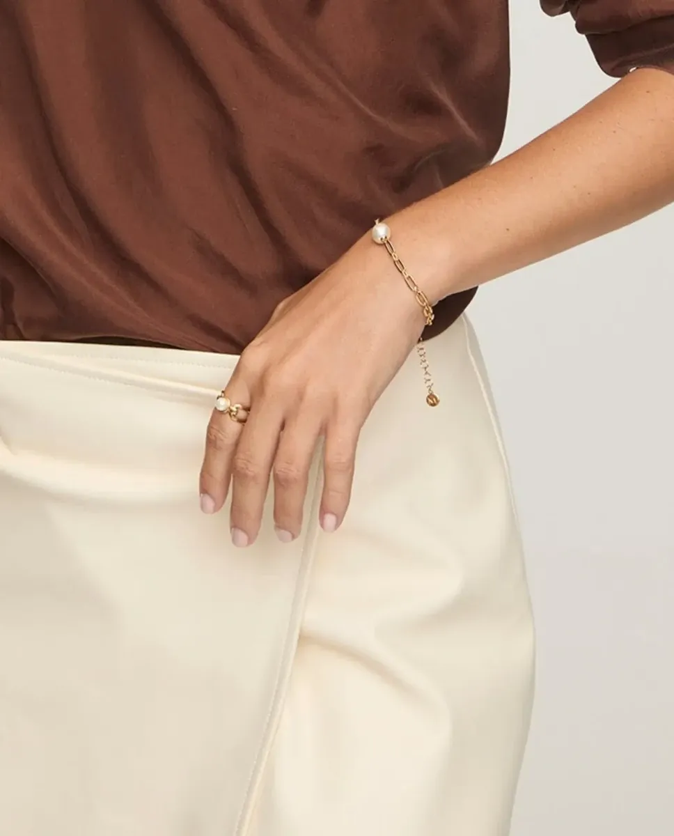 Main féminine portant une bague et un bracelet à maillons dorés et perles, sur fond de vêtements marron et blanc.