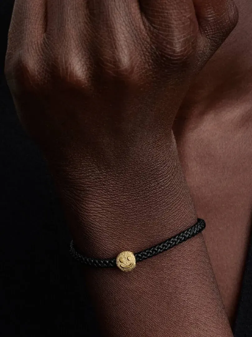 Close-up van de smiley charm aan een zwarte armband, gedragen om de pols van een model tegen een donkere achtergrond.