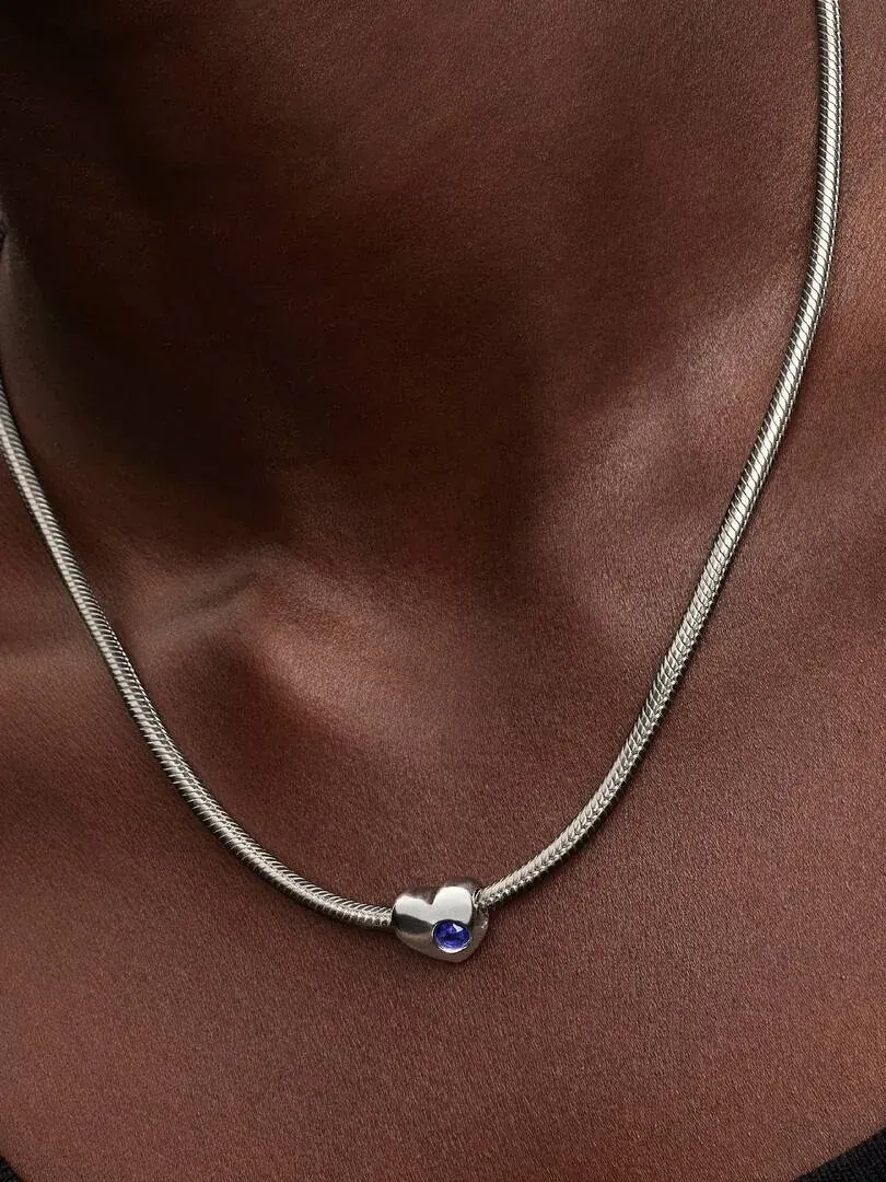 Primer plano del charm de corazón con cristal azul sobre una cadena de plata, puesto en el cuello.