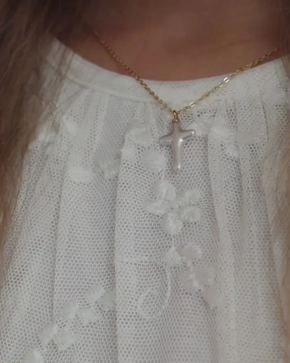 Primer plano del colgante de cruz sobre blusa blanca bordada, toma frontal en el cuello.