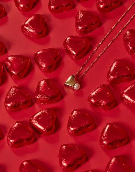 Collar con colgante de corazón dorado y perla sobre fondo rojo decorado con corazones metálicos brillantes.
