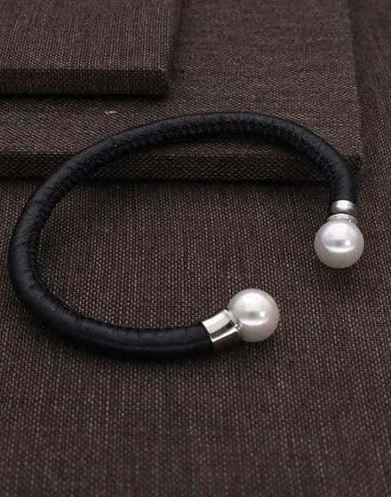 Zwarte leren armband met twee witte parels aan de uiteinden, bovenaanzicht op een bruine getextureerde achtergrond.
