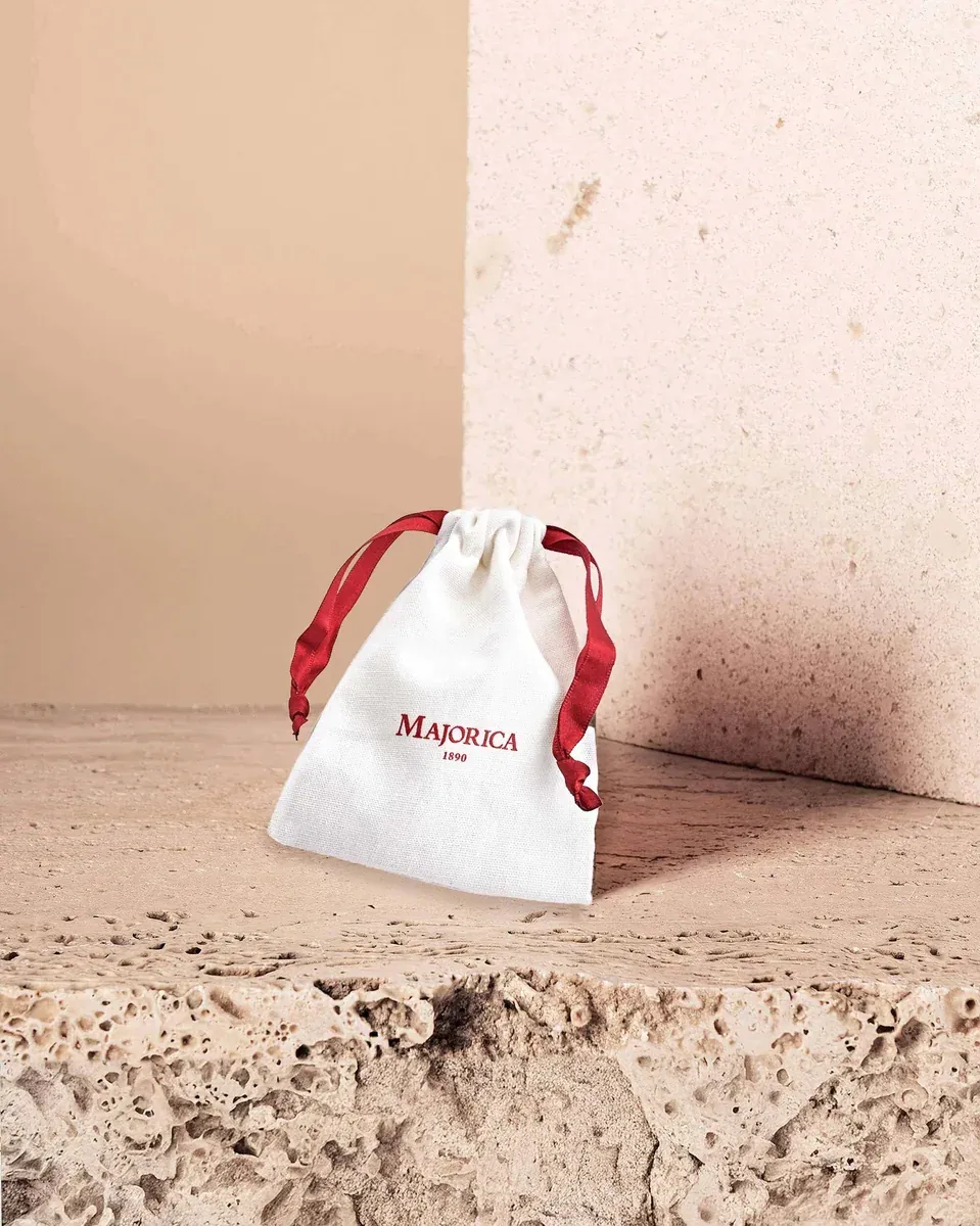 Bolsa de tela blanca con logotipo Majorica en rojo, sobre superficie de piedra natural texturizada.