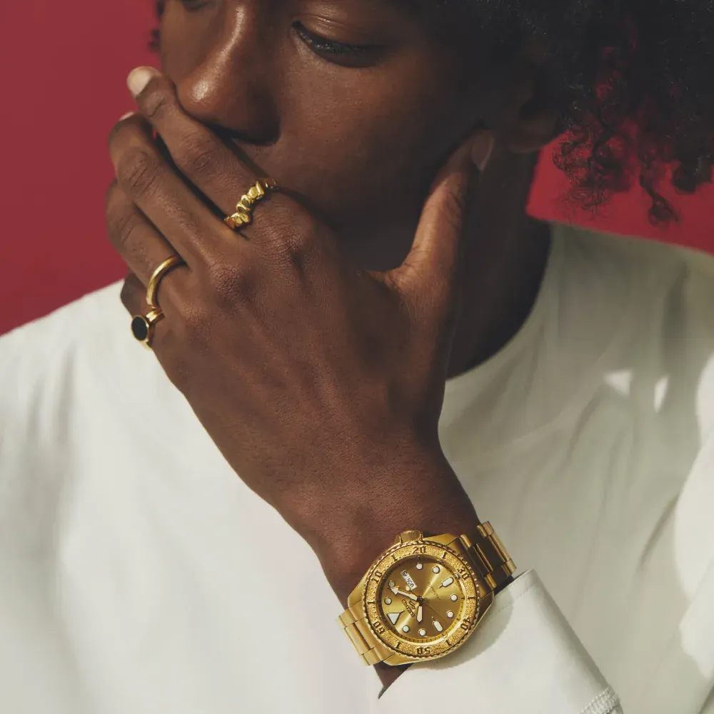 Modelo masculino con reloj dorado en la muñeca y anillos, camiseta blanca, fondo rojo