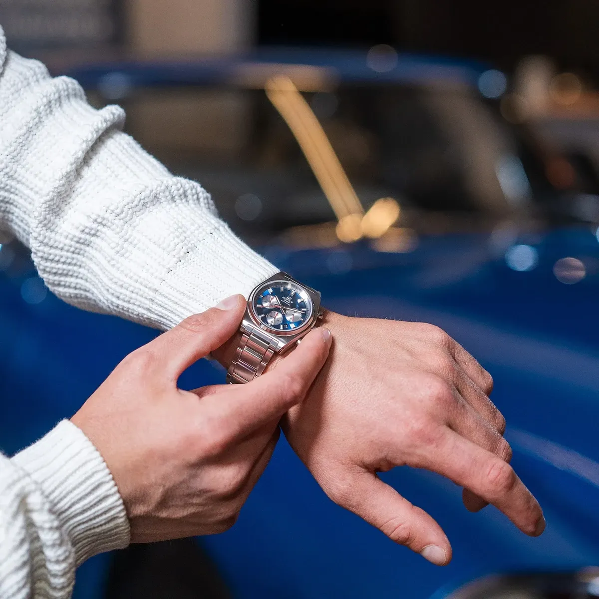 Reloj puesto en muñeca masculina con jersey blanco, fondo desenfocado de coche azul.