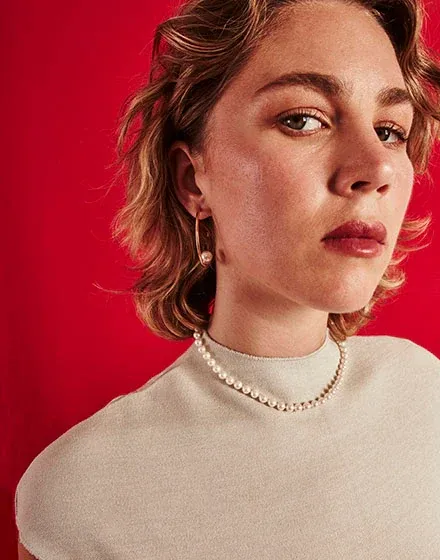 Nahaufnahme eines Models mit Perlenhalskette und Creolen vor lebhaft rotem Hintergrund.