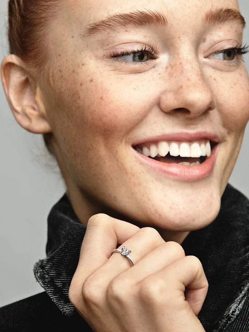Close-up of heart-shaped stone ring on finger, smiling model with neutral background.