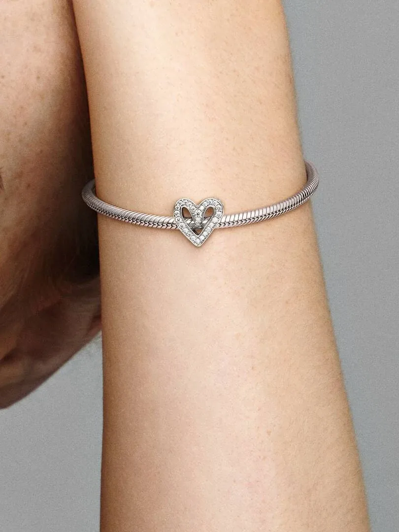 Close-up of the heart charm on a silver bracelet, worn on a wrist against a neutral background.