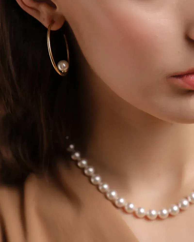 Primer plano del cuello y oreja de modelo con collar de perlas y pendiente de aro dorado, fondo neutro.