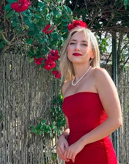 Model in rode jurk met parelsnoer, buitenopname met rode bloemen en een houten hek.
