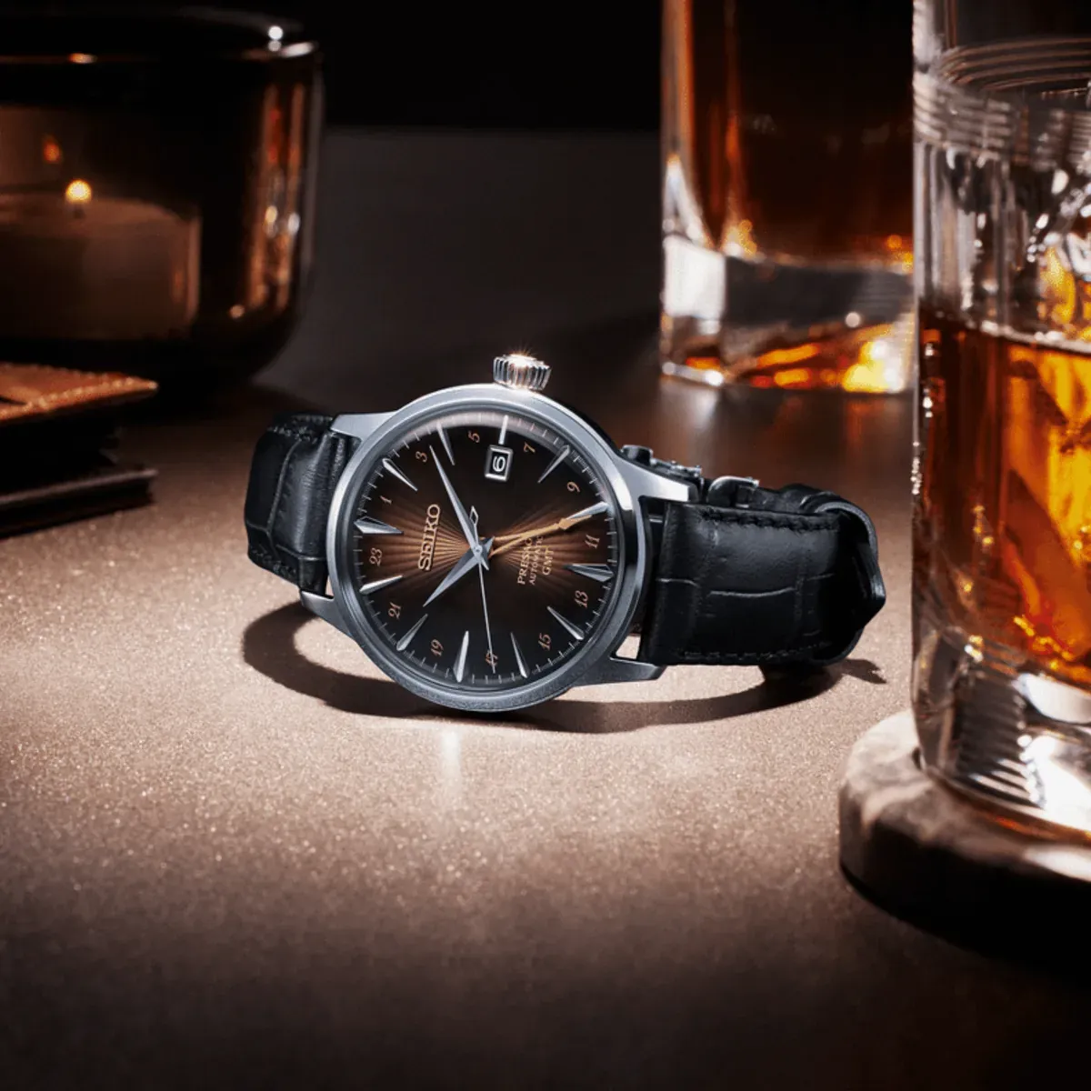 Reloj apoyado sobre superficie texturizada, composición con vasos de cóctel y vela al fondo, iluminación cálida.