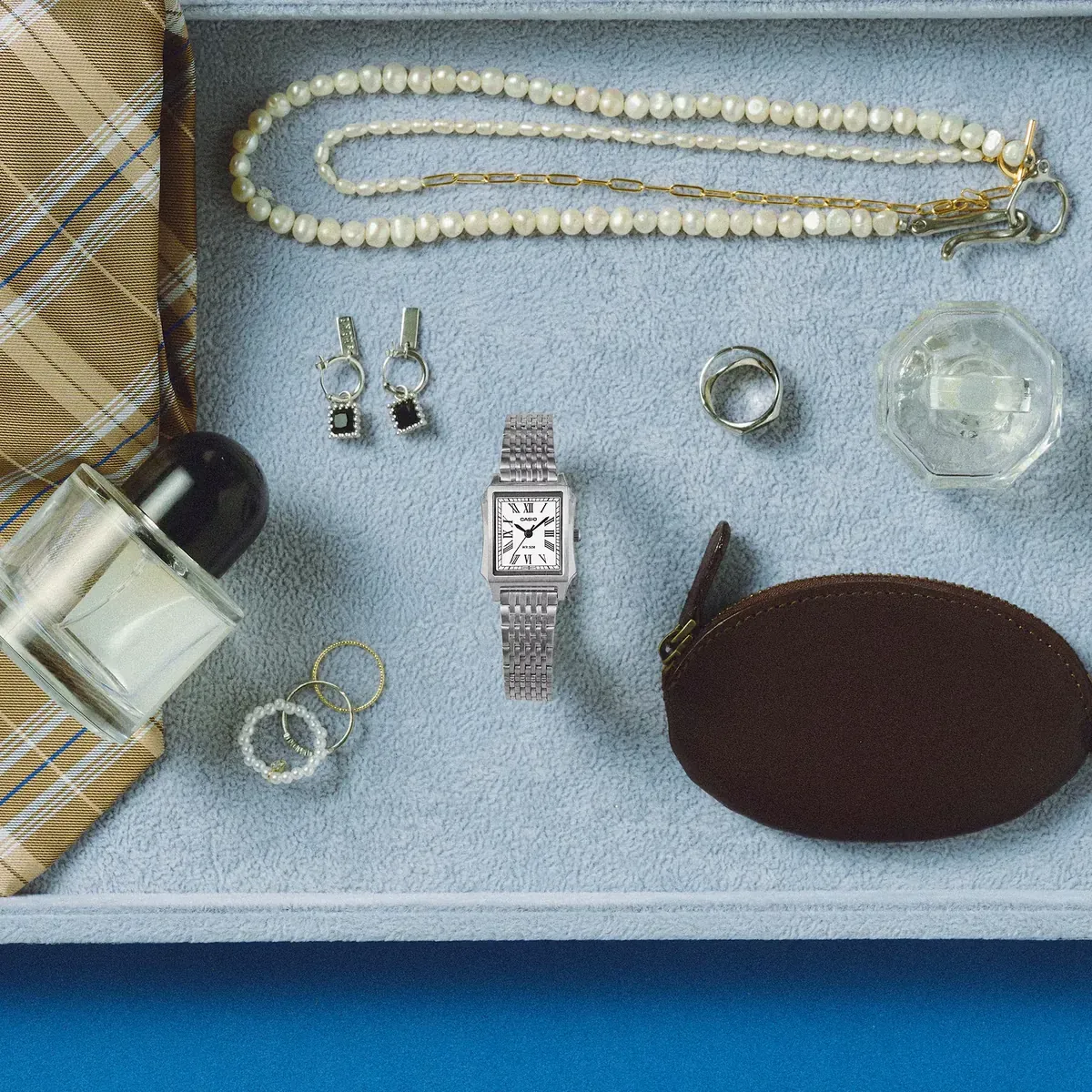 Vista cenital del reloj centrado en una bandeja gris junto a joyas, perfume y un estuche marrón.