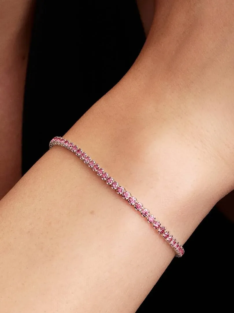 Close-up van de tennisarmband met roze stenen om een pols, diagonale opname tegen een donkere achtergrond.