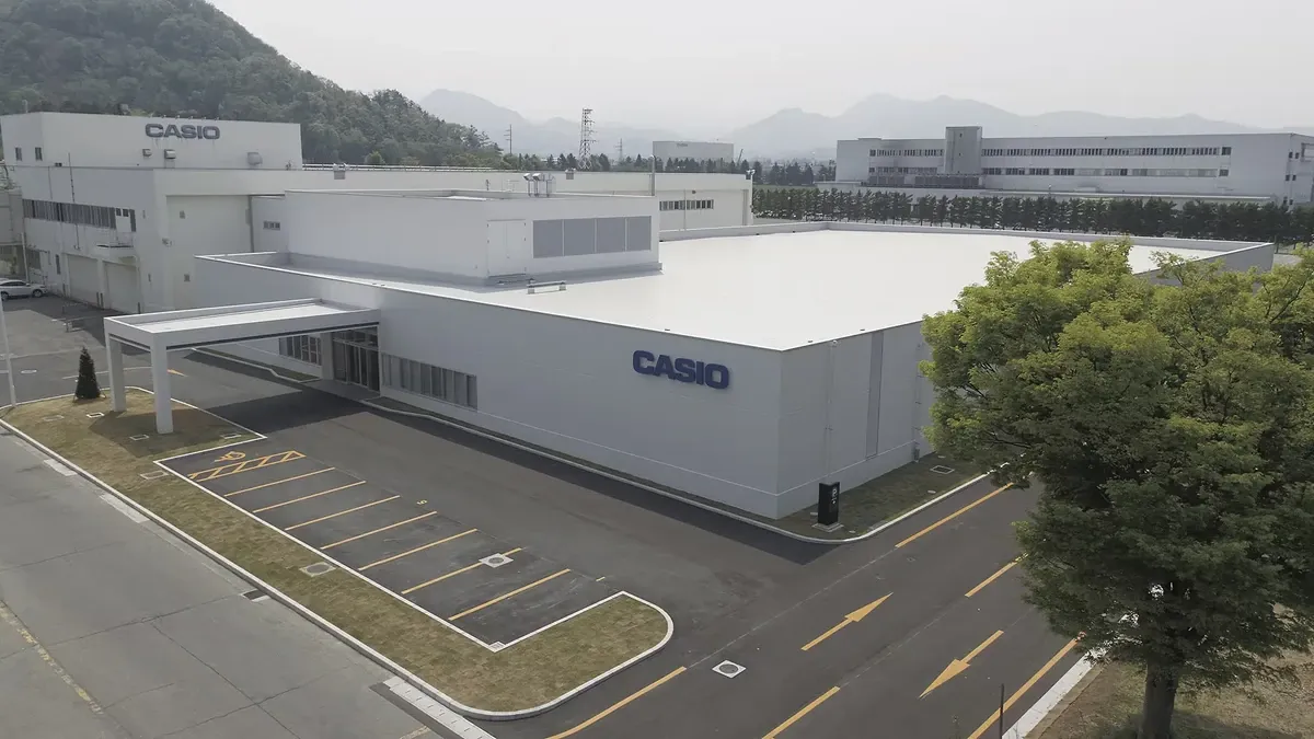 Vista aérea de las instalaciones industriales de Casio, con edificios blancos, aparcamiento y montañas al fondo.