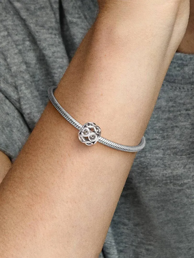 Close-up van een zilveren charm aan een snake chain armband, gedragen om de pols tegen een grijze achtergrond.