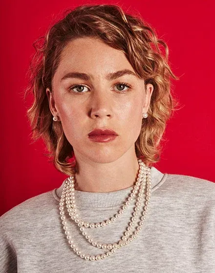 Nahaufnahme eines Models mit langer weißer Perlenkette über grauem Pullover, leuchtend roter Hintergrund.