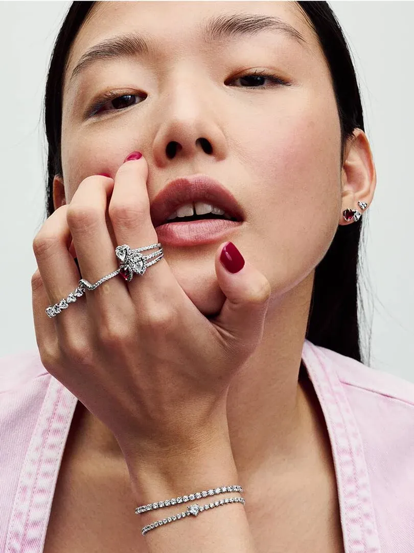 Close-up van een vrouwenhand met een hartring en een kubieke zirkonia armband tegen een neutrale achtergrond.