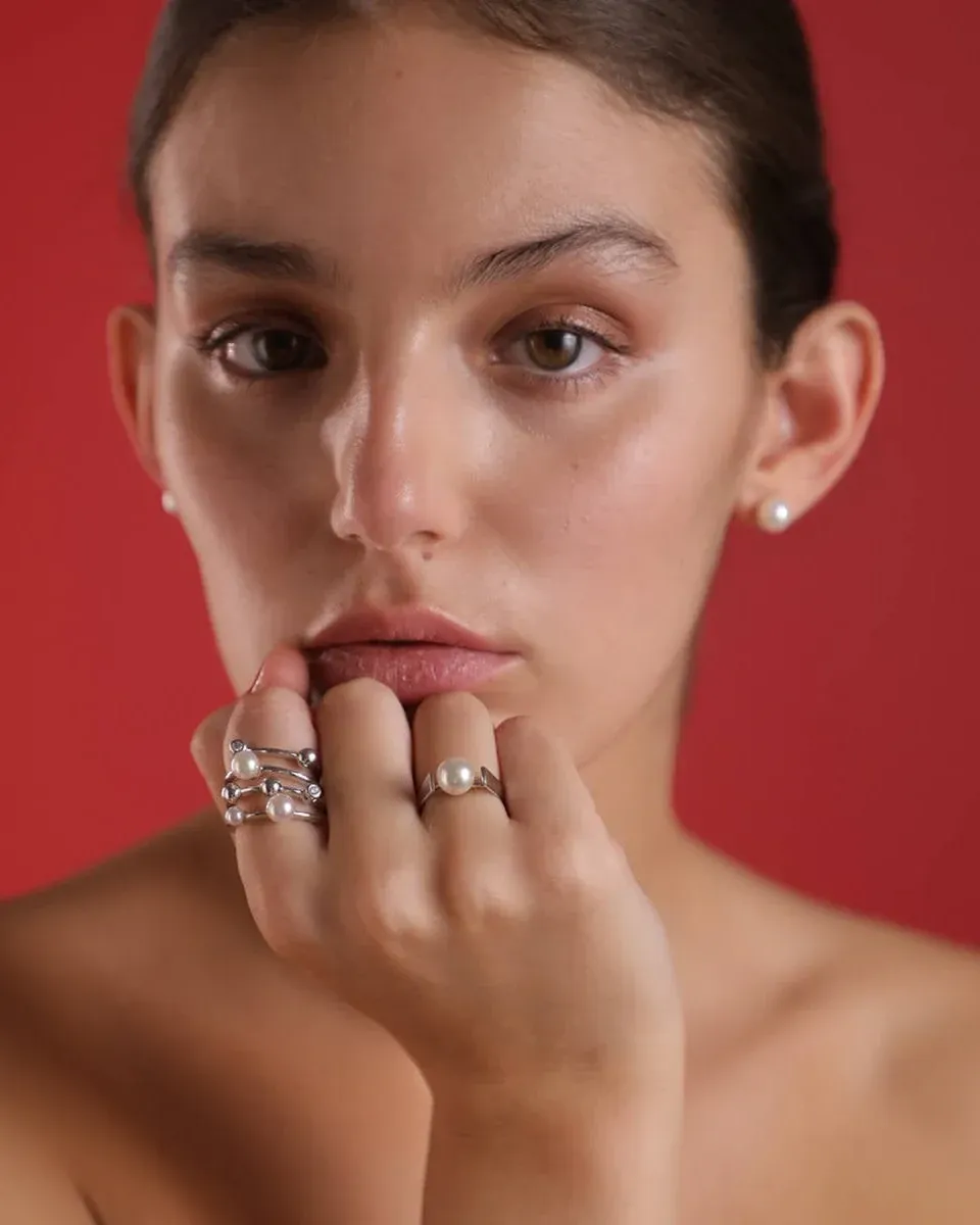 Close-up van een hand met een witte parelring, model tegen een effen rode achtergrond.