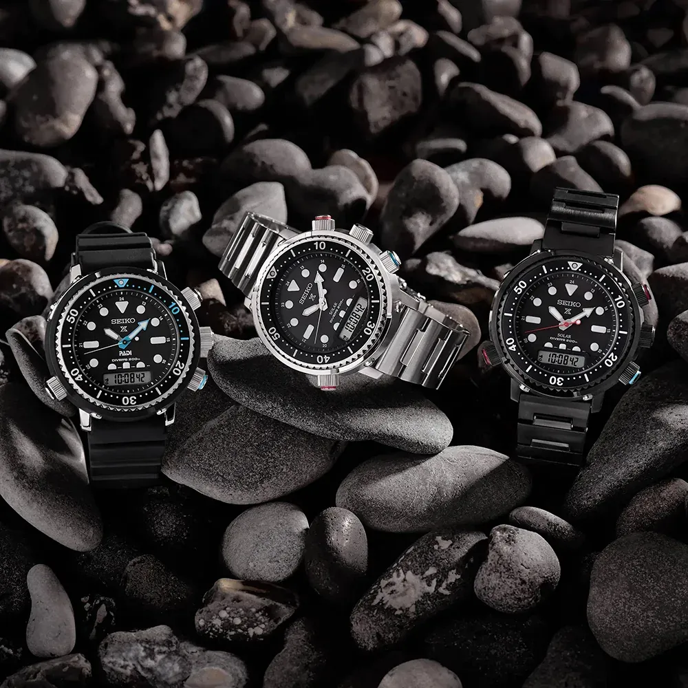 Tres variantes del reloj dispuestas sobre piedras oscuras y lisas, vista cenital.