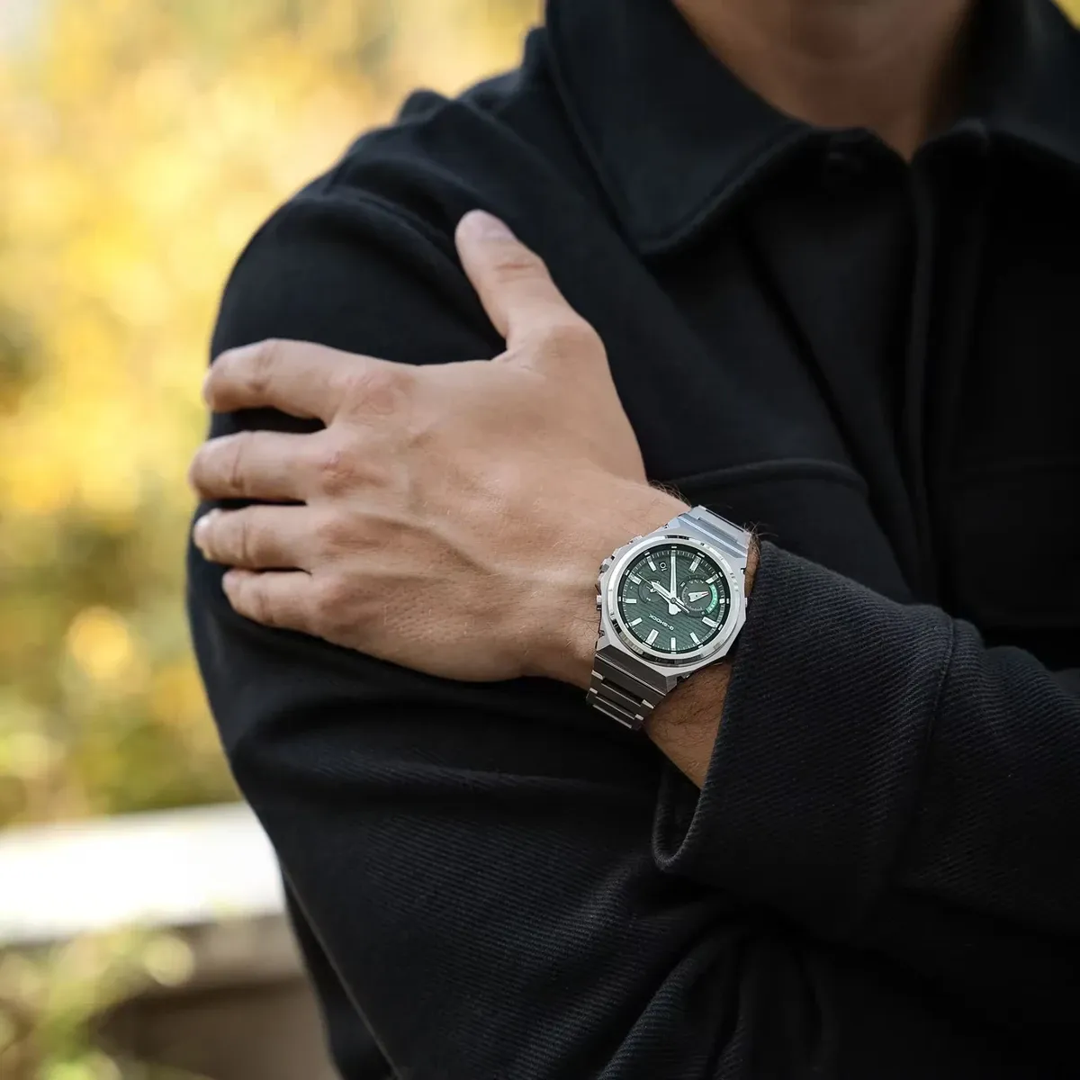 Primer plano del reloj puesto en la muñeca, modelo con prenda negra y fondo otoñal desenfocado.